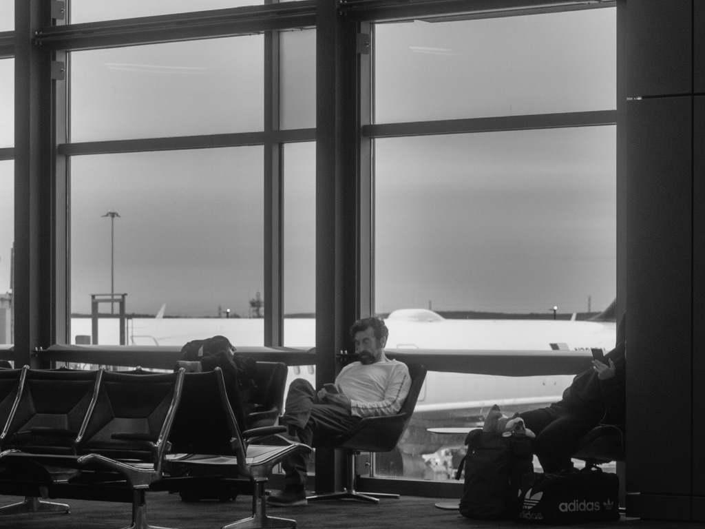 A man waits alone in an airport terminal, viewing airplanes through large windows on an overcast day.