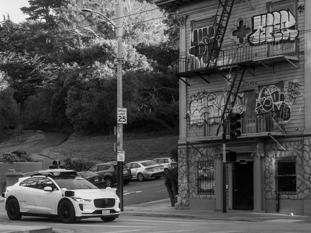 A white electric vehicle parked on an urban street corner next to a graffitied building with trees overhead.