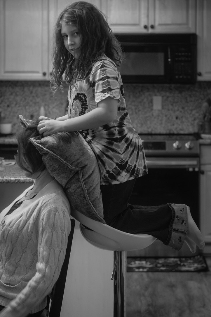 A young girl sits on a kitchen counter wearing tie-dye, looking back over her shoulder with a playful expression.