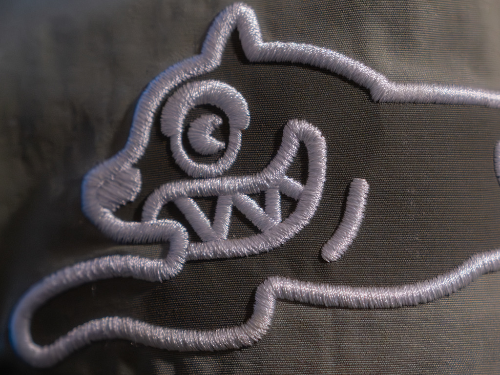 Close-up of silver embroidered decorative flourish with scrolling ornamental details on gray fabric.