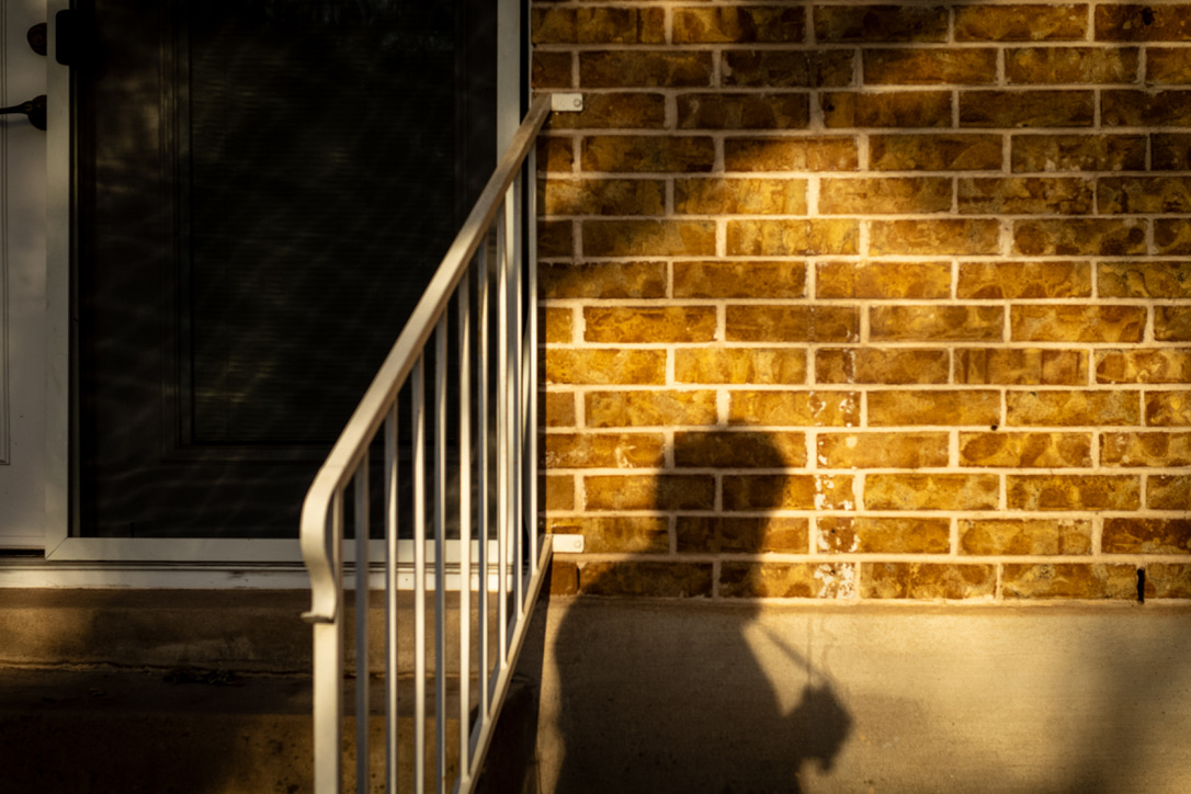 Sunlit brick wall and metal railing cast sharp shadows on a concrete surface at dusk or evening.