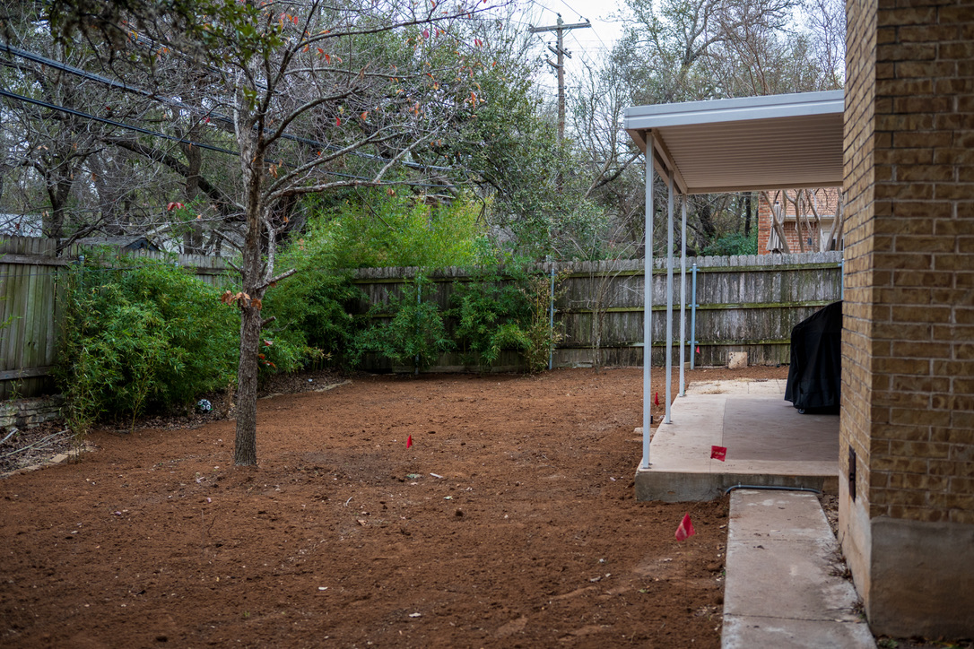 A backyard under construction with a brick house, metal carport, bare soil, and surveyor's flags marking a landscaping or building project.