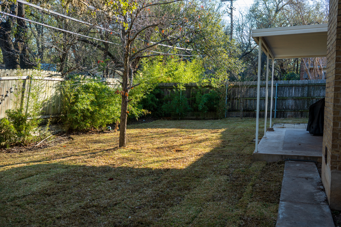 A sunny backyard with mature trees, patchy grass, wooden fences, and a brick house with a covered patio structure.