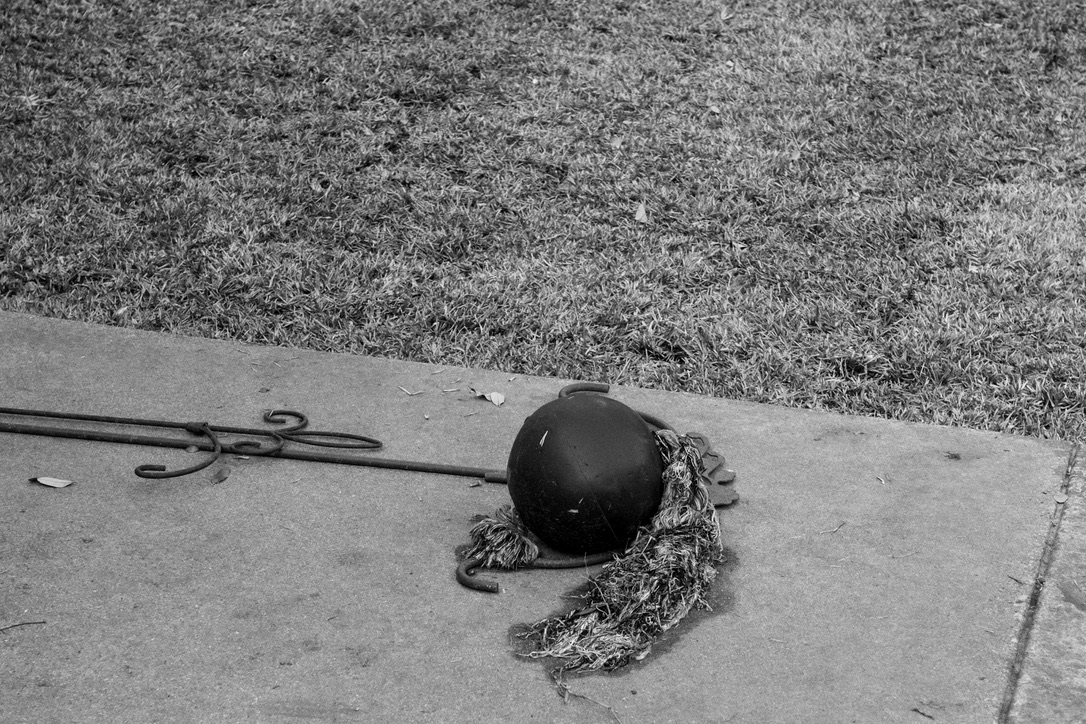 A black buoy with tangled rope and seaweed lies on a concrete surface beside dry grass in black and white.