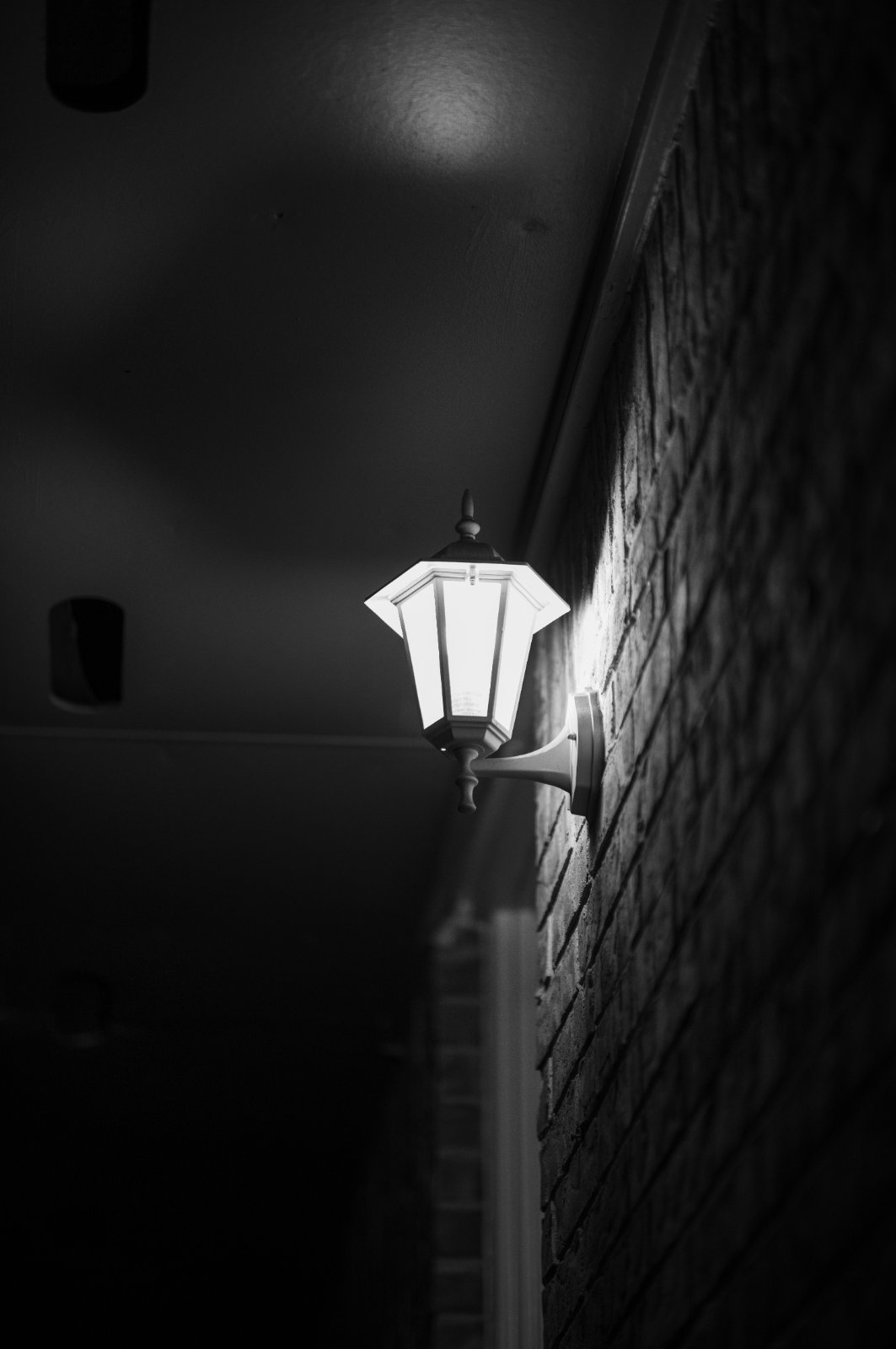 A solitary illuminated street lamp glows against a dark brick wall in a moody nighttime alley.
