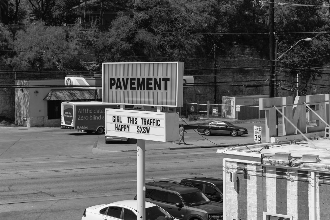 Black and white street scene with humorous traffic sign reading 'Girl This Traffic Happy SXSW' amid urban construction and parked cars.