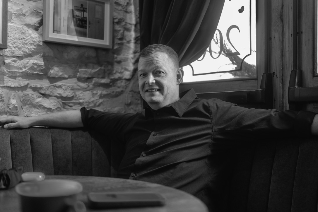 A man in a dark shirt sits relaxed on a banquette in a rustic stone-walled establishment, smiling at the camera.