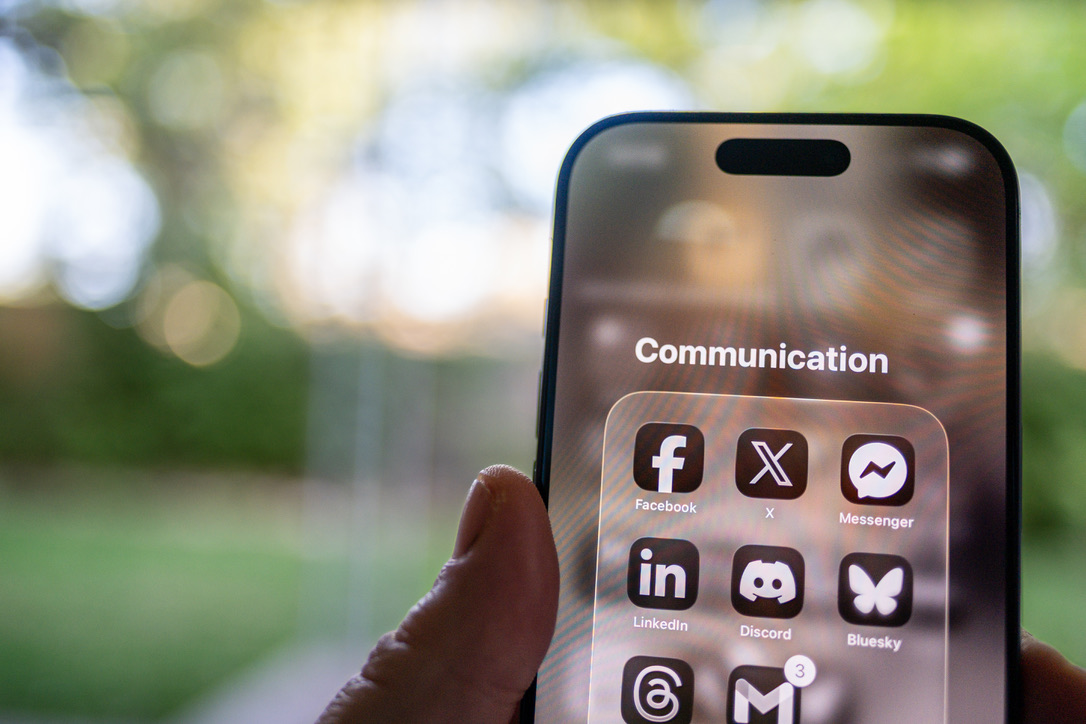A hand holding a smartphone displaying a Communication folder with social media and messaging app icons against a blurred outdoor background.