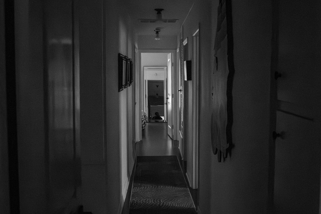 A narrow, dimly-lit hallway with white walls and open doors stretches into the distance with a figure visible at the far end.