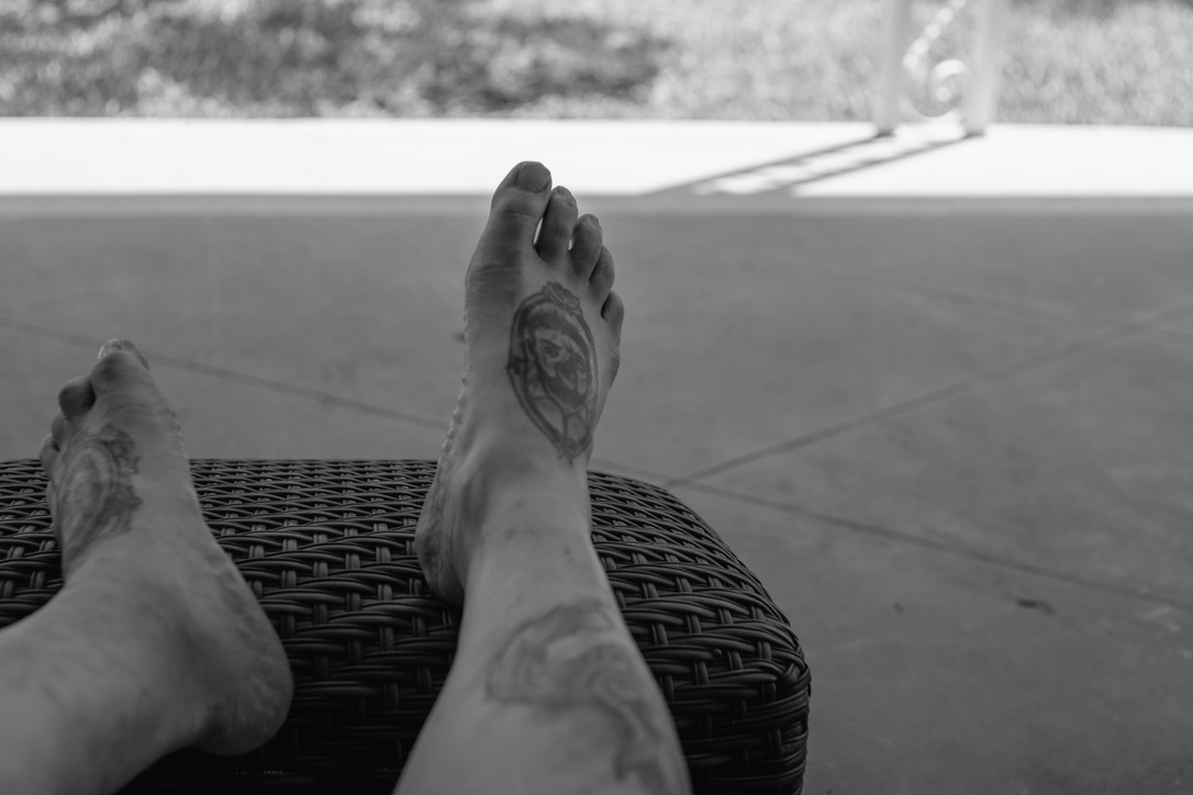 Bare feet propped on a woven chair overlooking a calm lake or water body with blurred greenery in the background.