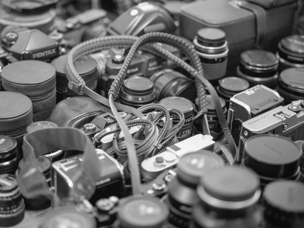 A collection of vintage film cameras and lenses with a braided camera strap draped across the gear in black and white.