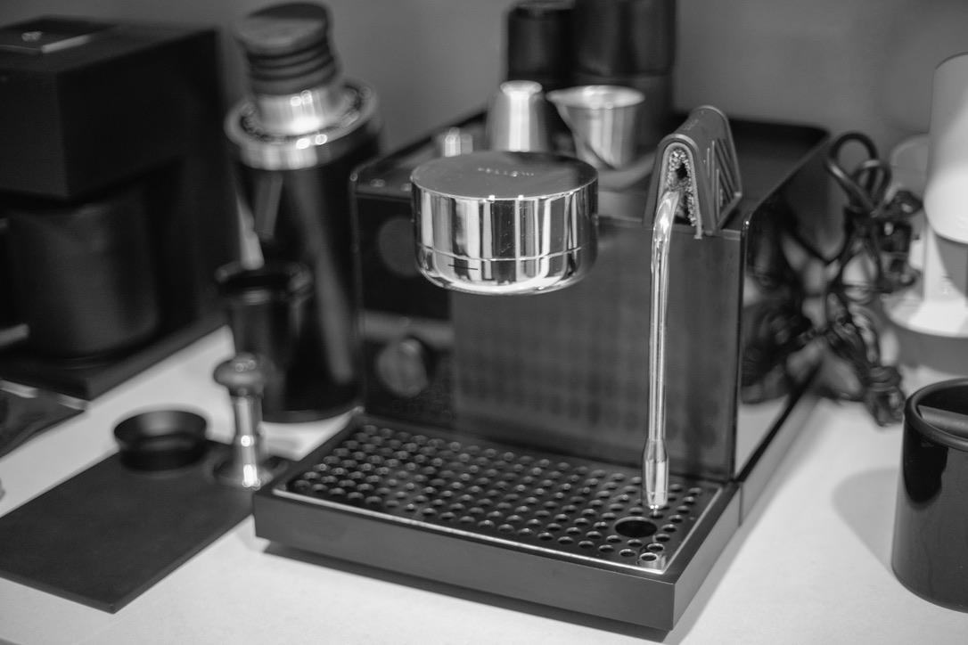 A monochrome overhead view of a professional espresso machine setup with various metal containers and tools on a white counter.